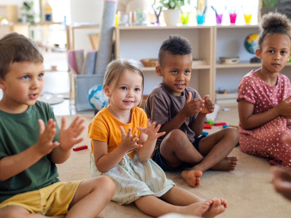 Group of small nursery school children sitting on floor indoors in classroom, clapping.